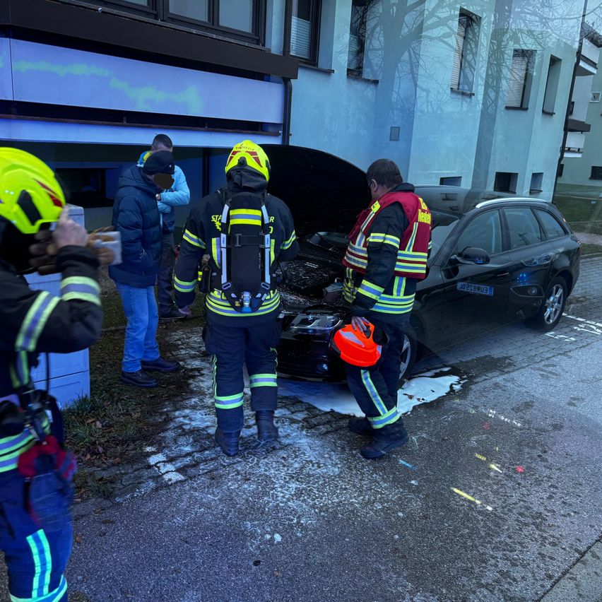 Feuerwehrleute inspizieren ein demoliertes Auto vor einem Gebäude. Mehrere Personen in Schutzkleidung stehen in der Nähe. Eine Person hält einen Becher.