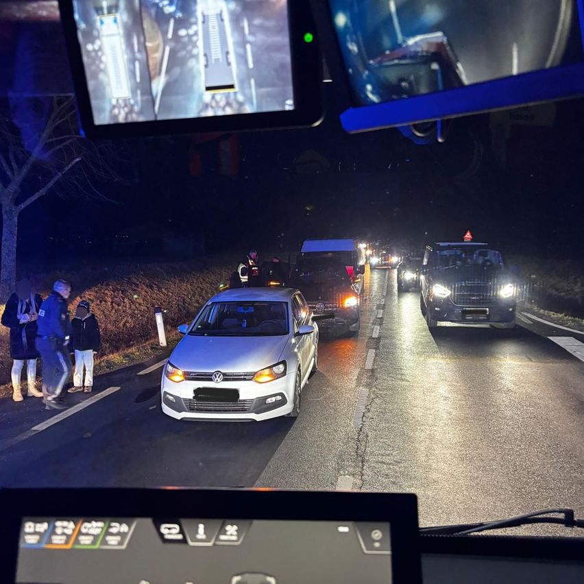 Ein Konvoi von Fahrzeugen fährt bei Nacht auf einer Straße, mit einem Volkswagen im Vordergrund. Einige Menschen stehen am Straßenrand, einer trägt eine Warnweste.