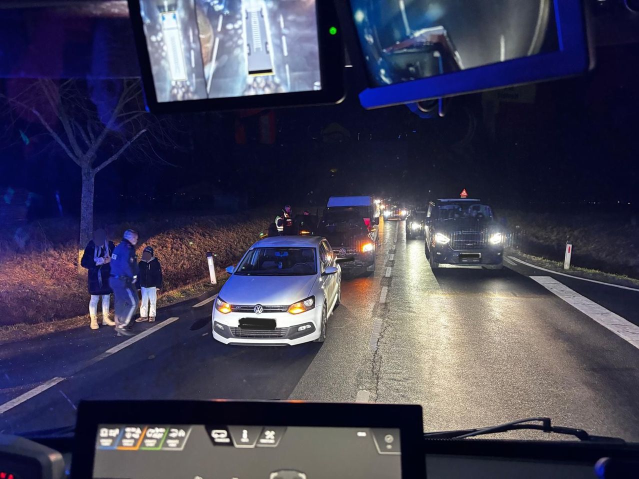 Ein Konvoi von Fahrzeugen fährt bei Nacht auf einer Straße, mit einem Volkswagen im Vordergrund. Einige Menschen stehen am Straßenrand, einer trägt eine Warnweste.