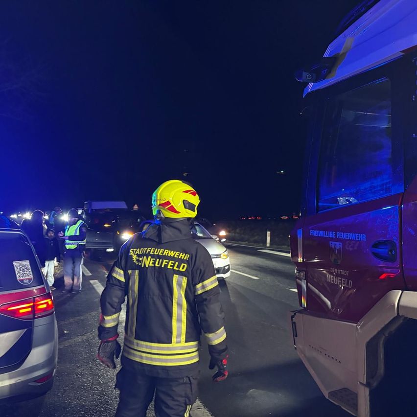 Ein Feuerwehrmann der Freiwilligen Feuerwehr Neufeld steht bei Nacht an einer Straße. In der Nähe befinden sich mehrere Personen und Fahrzeuge. Der Feuerwehrmann trägt einen gelben Helm und eine reflektierende Weste. Ein roter Feuerwehrwagen ist in der Nähe geparkt.