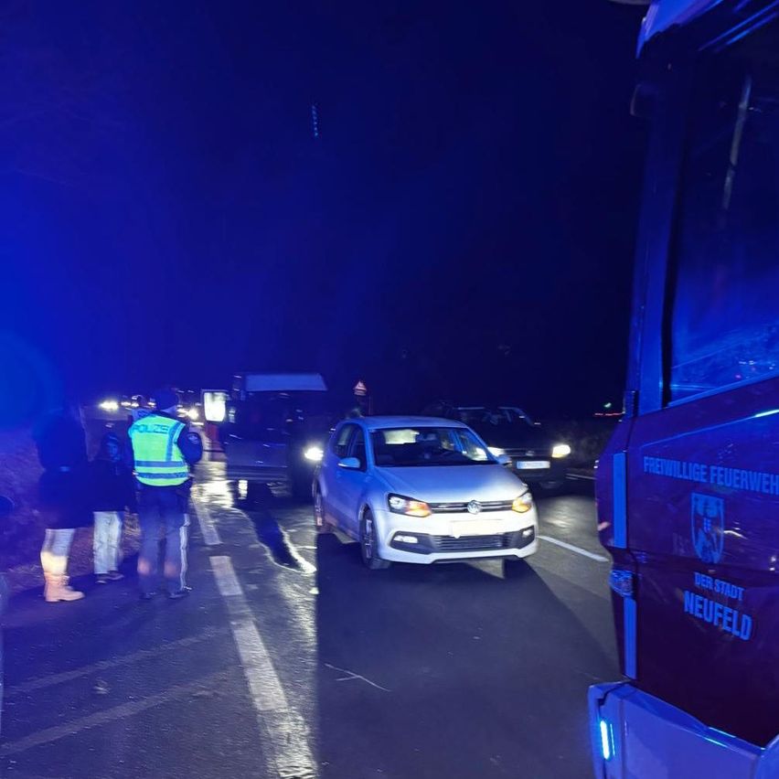 In der Nacht wird ein weißer Wagen von einem Polizisten angehalten. Der Polizist trägt eine gelbe Weste und einen Hut. Der Wagen ist am Straßenrand geparkt.