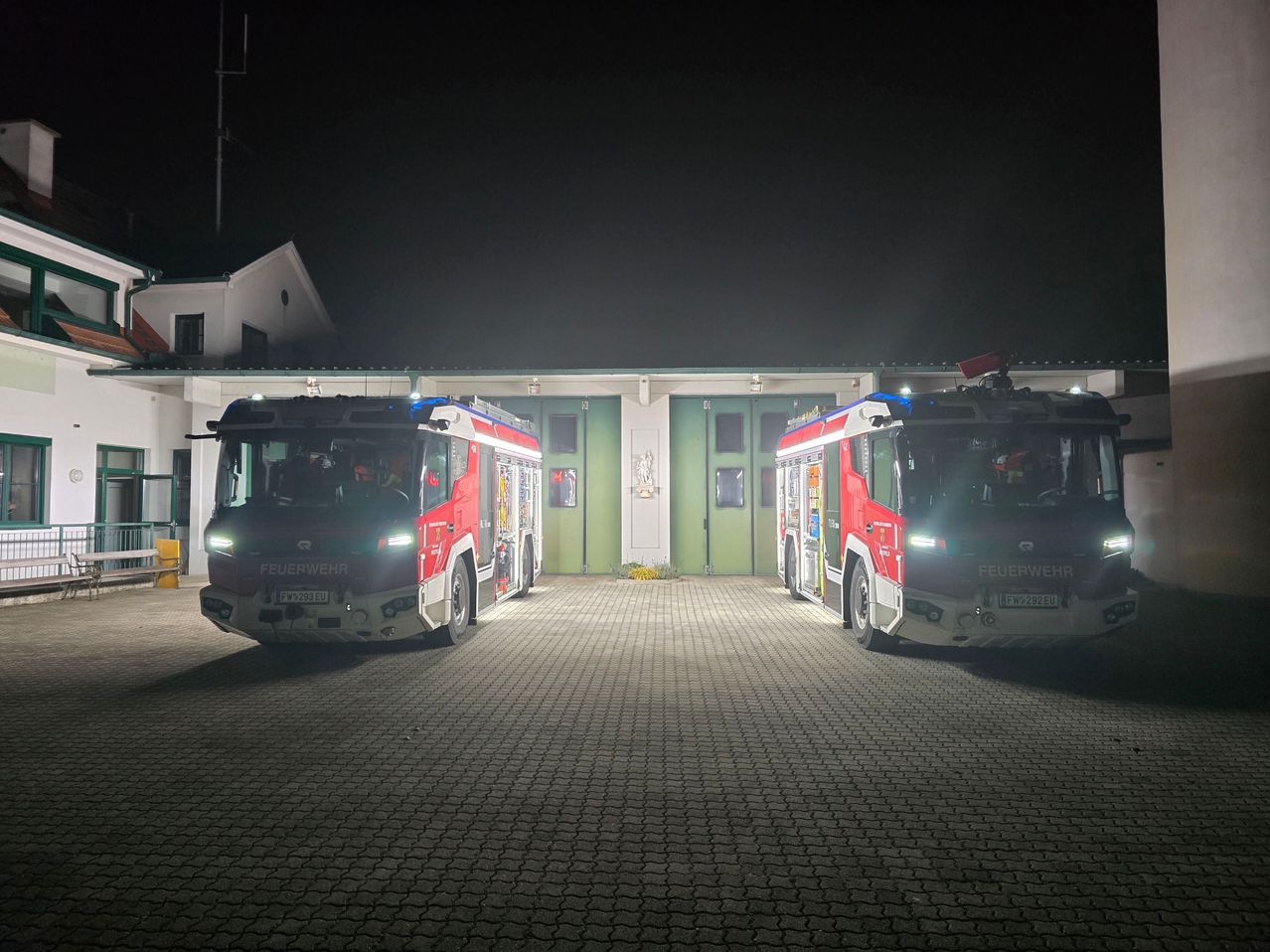 Zwei Feuerwehrfahrzeuge stehen nachts vor einer Feuerwache. Die Feuerwehrfahrzeuge haben Lichter eingeschaltet und sind einsatzbereit.