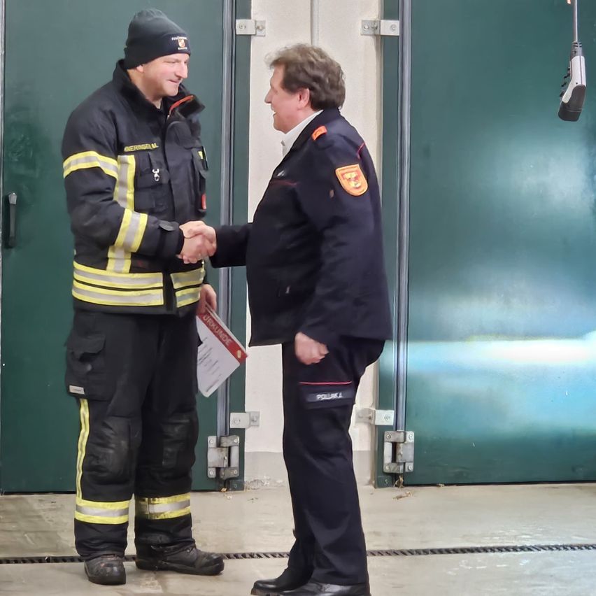 Zwei Männer, einer in Feuerwehruniform und der andere in formeller Kleidung, schütteln sich die Hände. Sie stehen vor einer grünen Tür.