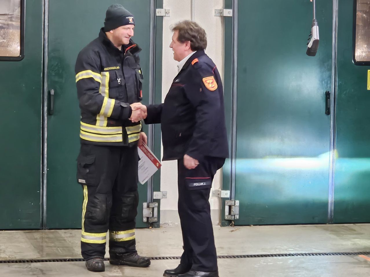 Zwei Männer, einer in Feuerwehruniform und der andere in formeller Kleidung, schütteln sich die Hände. Sie stehen vor einer grünen Tür.