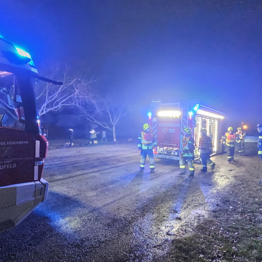 Feuerwehrleute in Uniformen sind auf einer Straße bei Nacht, mit einem Feuerwehrwagen und blinkenden Lichtern.