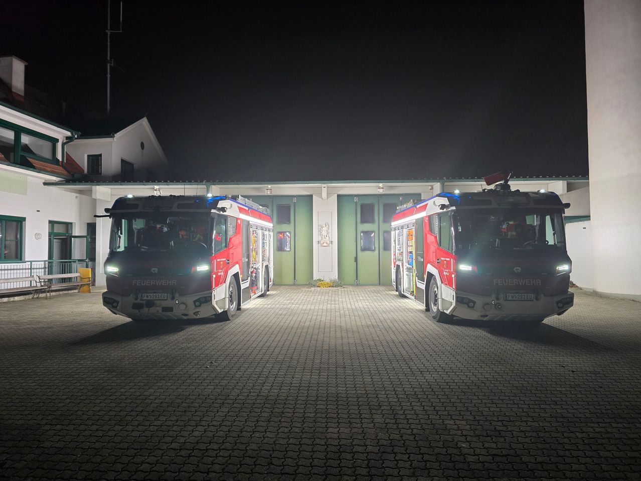 Zwei Feuerwehrfahrzeuge sind nachts mit eingeschalteten Scheinwerfern in einer Garage geparkt.