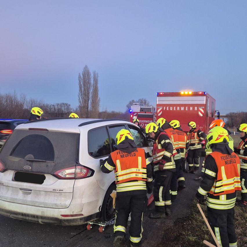 Feuerwehrleute in leuchtend gelben Helmen und Schutzanzügen kümmern sich um ein beschädigtes weißes Auto. Der Ort der Szene ist eine Straße mit einem roten Feuerwehrwagen im Hintergrund.