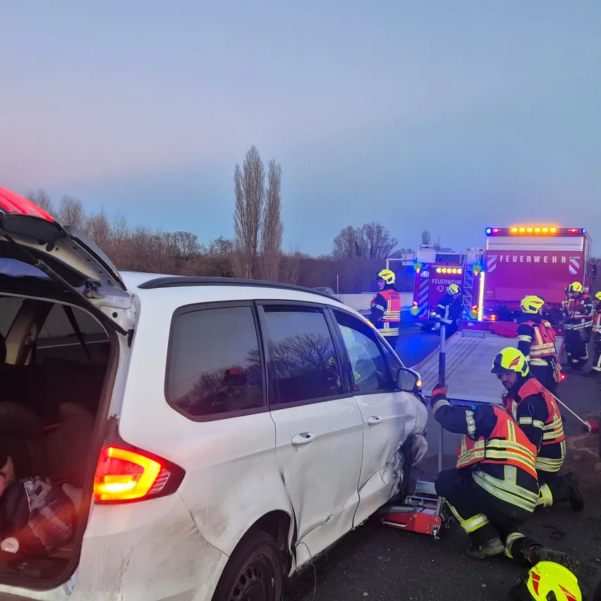 Rettungskräfte kümmern sich um ein beschädigtes weißes Auto am Straßenrand. Ein Feuerwehrmann benutzt einen hydraulischen Wagenheber, um das Auto anzuheben.