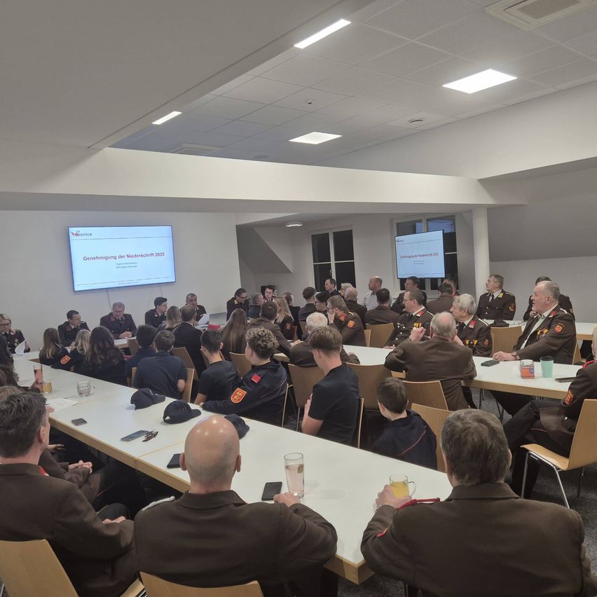 Eine Versammlung in einem Konferenzraum mit Teilnehmern an Tischen. Eine Beamerleinwand zeigt eine Präsentation. Die Teilnehmer tragen Uniformen, einige mit Abzeichen. Der Raum hat weiße Wände, Deckenleuchten und Fenster.