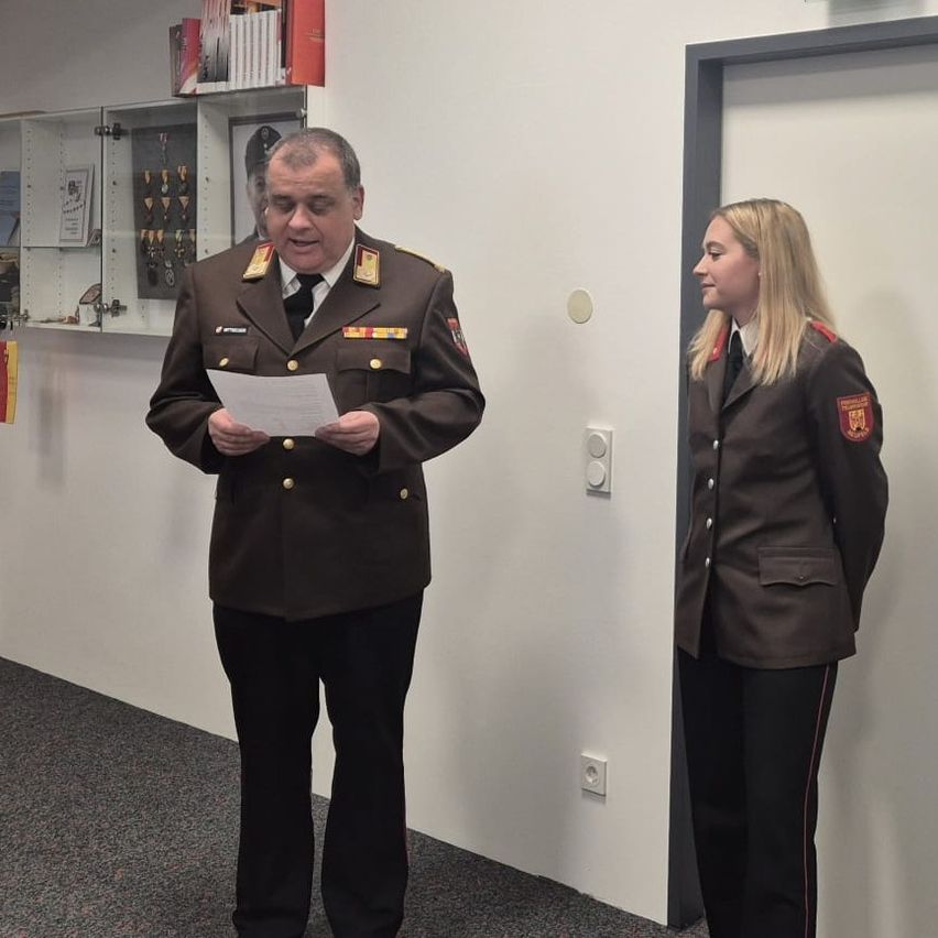 Ein Mann in Uniform steht und liest ein Papier, während eine Frau in Uniform zuhört. Hinter ihnen hat eine weiße Wand Regale mit Medaillen und Büchern.