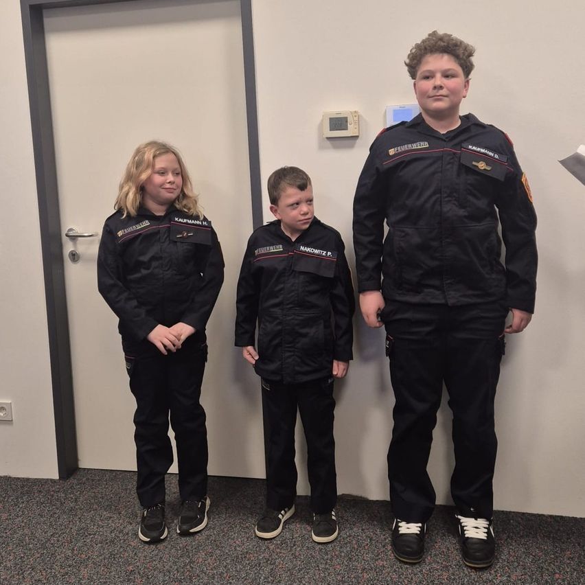 Drei Kinder in schwarzen Uniformen stehen nebeneinander in einem Raum. Sie tragen Turnschuhe und scheinen zu lächeln.