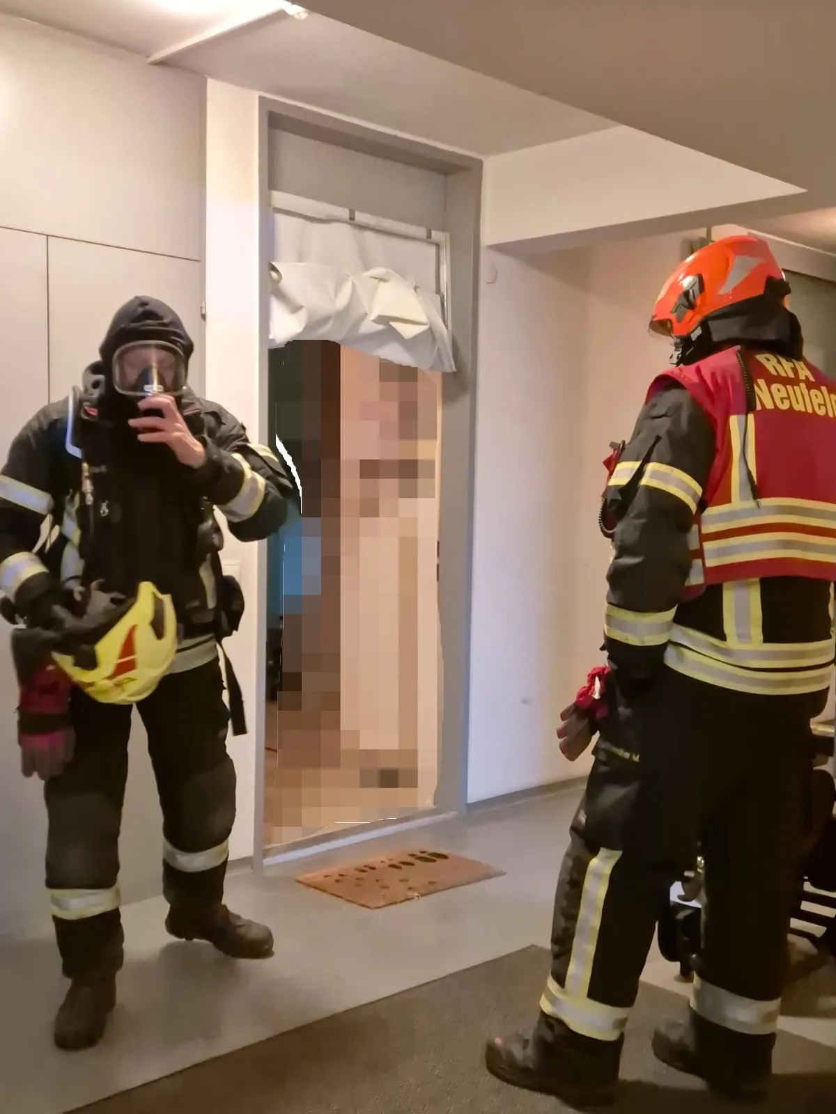 Zwei Feuerwehrleute in voller Ausrüstung, einer mit Maske, stehen in einem Raum mit offener Tür. Ein verschwommenes Bild einer Person ist im Hintergrund zu sehen.