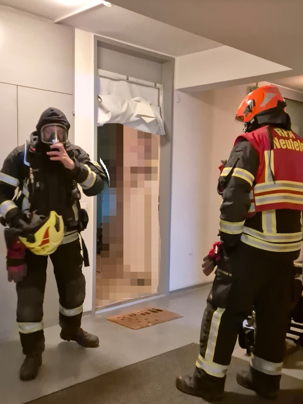Zwei Feuerwehrleute in voller Ausrüstung, einer mit Maske, stehen in einem Raum mit offener Tür. Ein verschwommenes Bild einer Person ist im Hintergrund zu sehen.