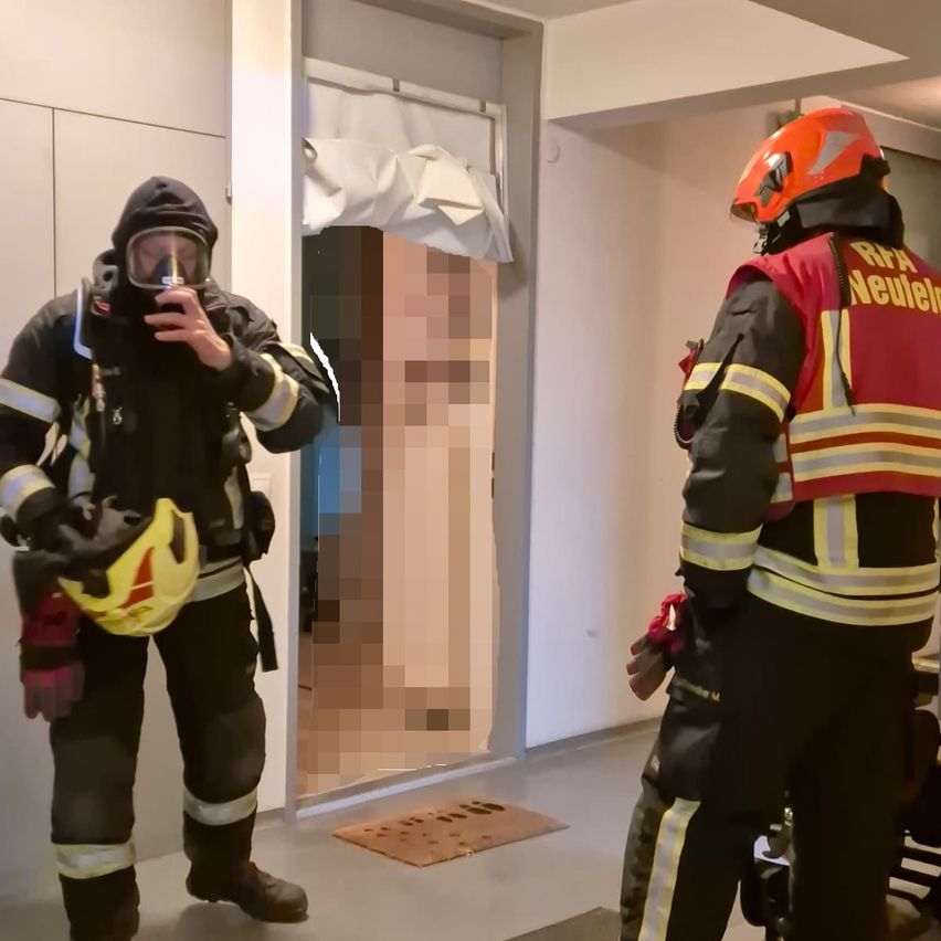 Zwei Feuerwehrleute in voller Ausrüstung, einer mit Maske, stehen in einem Raum mit offener Tür. Ein verschwommenes Bild einer Person ist im Hintergrund zu sehen.