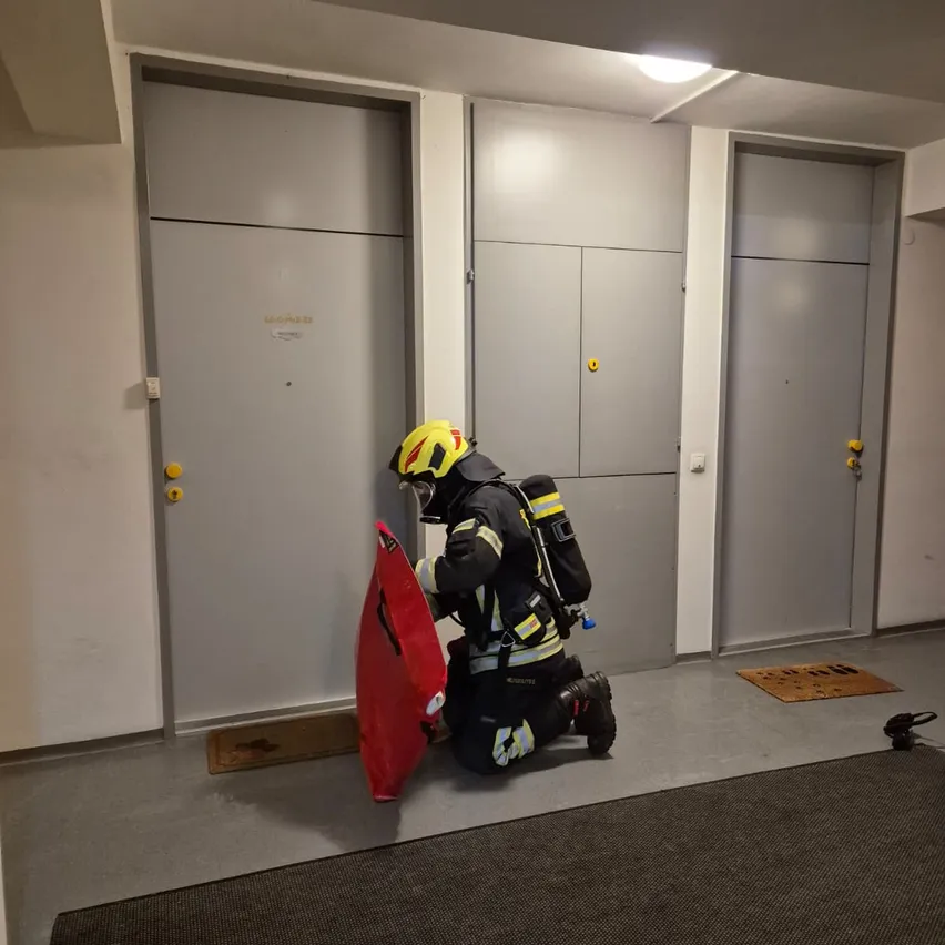 Ein Feuerwehrmann mit Helm und Schutzausrüstung kniet in einem Flur und hält einen roten Schild. Hinter ihm befinden sich drei geschlossene graue Türen und ein grauer Teppichboden.