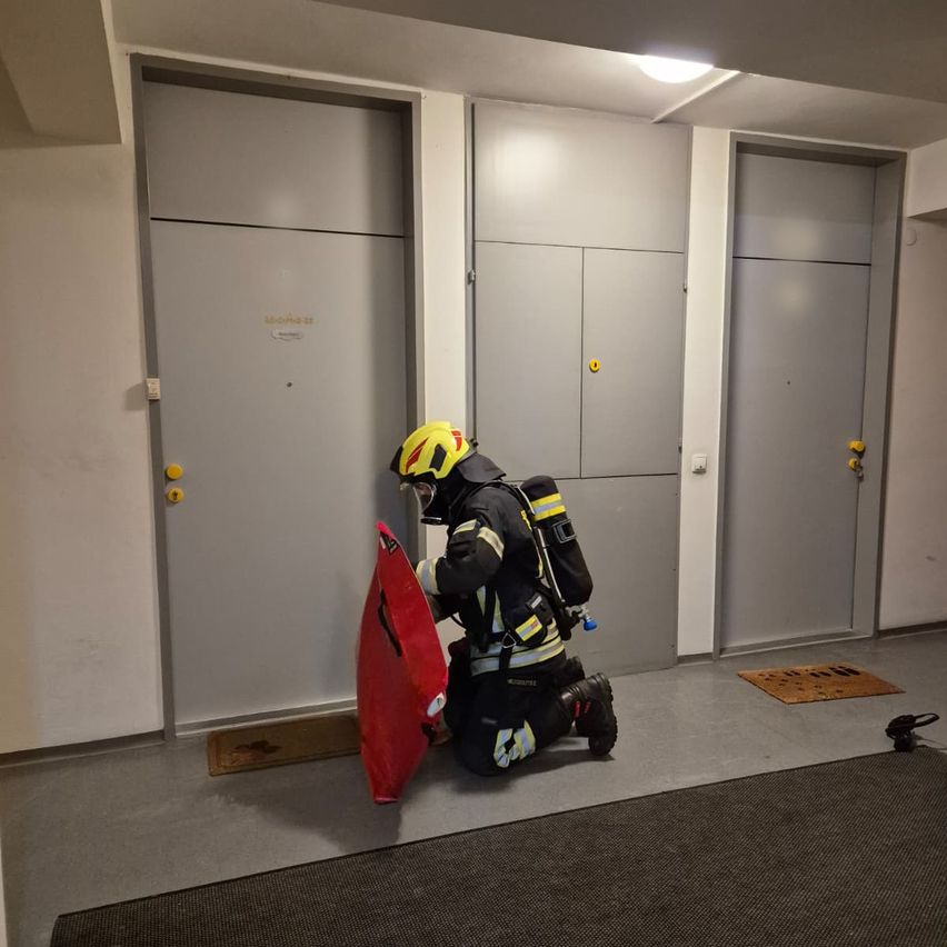 Ein Feuerwehrmann mit Helm und Schutzausrüstung kniet in einem Flur und hält einen roten Schild. Hinter ihm befinden sich drei geschlossene graue Türen und ein grauer Teppichboden.