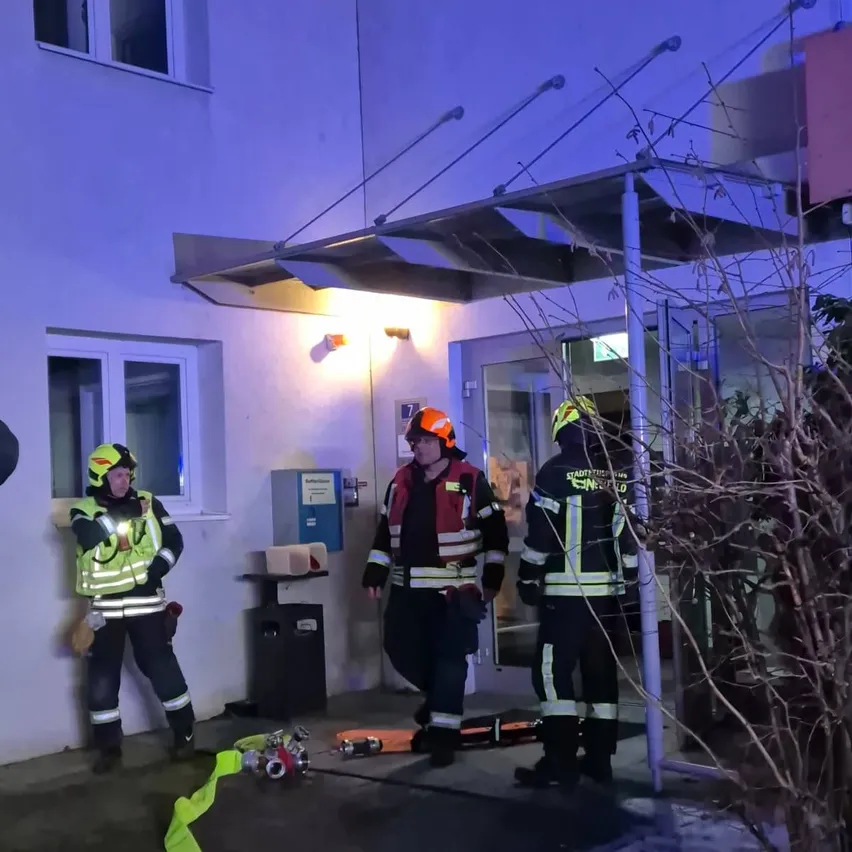 Drei Feuerwehrleute in Schutzkleidung stehen vor einem Gebäude, einer hält eine Taschenlampe, ein anderer einen Feuerlöscher und ein Dritter steht in der Nähe einer offenen Tür.