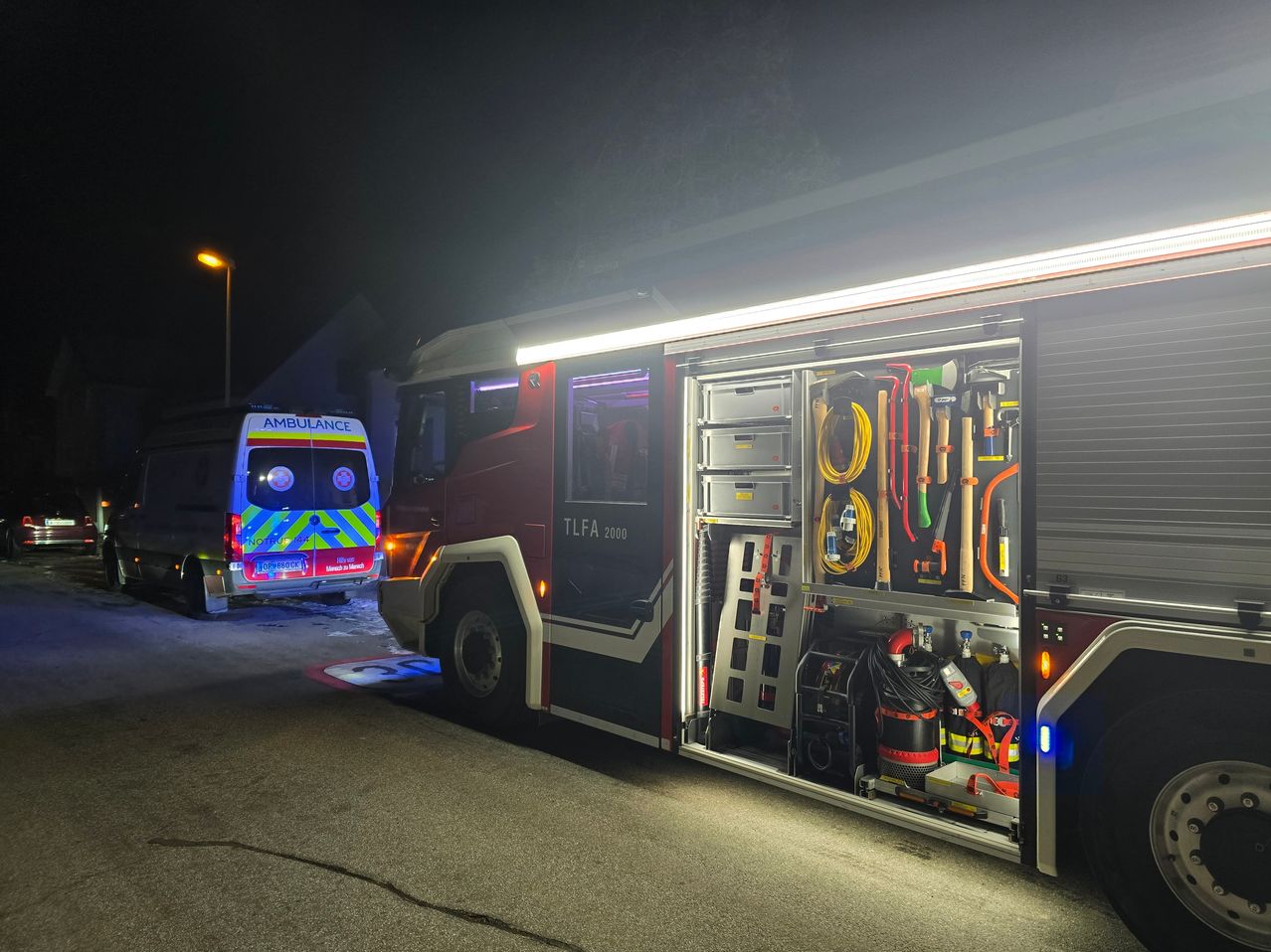 Ein Feuerwehrauto ist geparkt, mit offenen Türen, die die Innenausstattung zeigen. Ein Krankenwagen ist im Dunkeln dahinter geparkt.