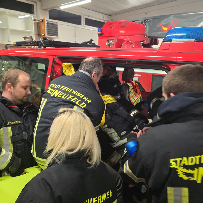 Feuerwehrleute stehen um einen roten Feuerwehrwagen mit eingeschalteten Notfalllichtern herum. Einige tragen Warnwesten. Eine Frau vorne hat blonde Haare.