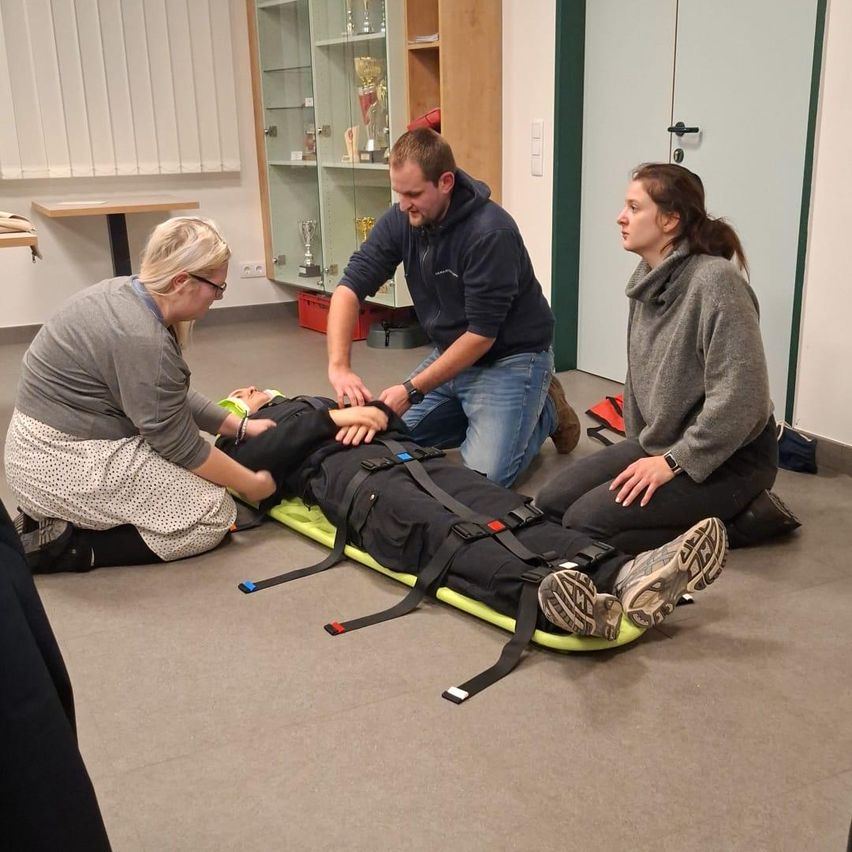 Drei Personen sind in einem Raum, eine Person liegt auf einer Trage, während die anderen beiden Hilfe leisten. Die Person auf der Trage trägt einen Helm und Turnschuhe. Hinter ihnen steht ein Glasregal mit Trophäen.