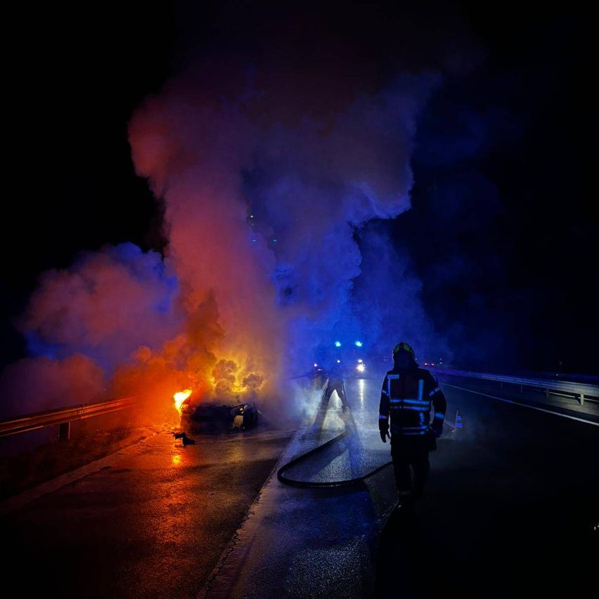 Ein Feuerwehrmann geht auf eine brennende Karre auf einer nassen Straße bei Nacht zu, mit Rauch und blinkenden Lichtern im Hintergrund.