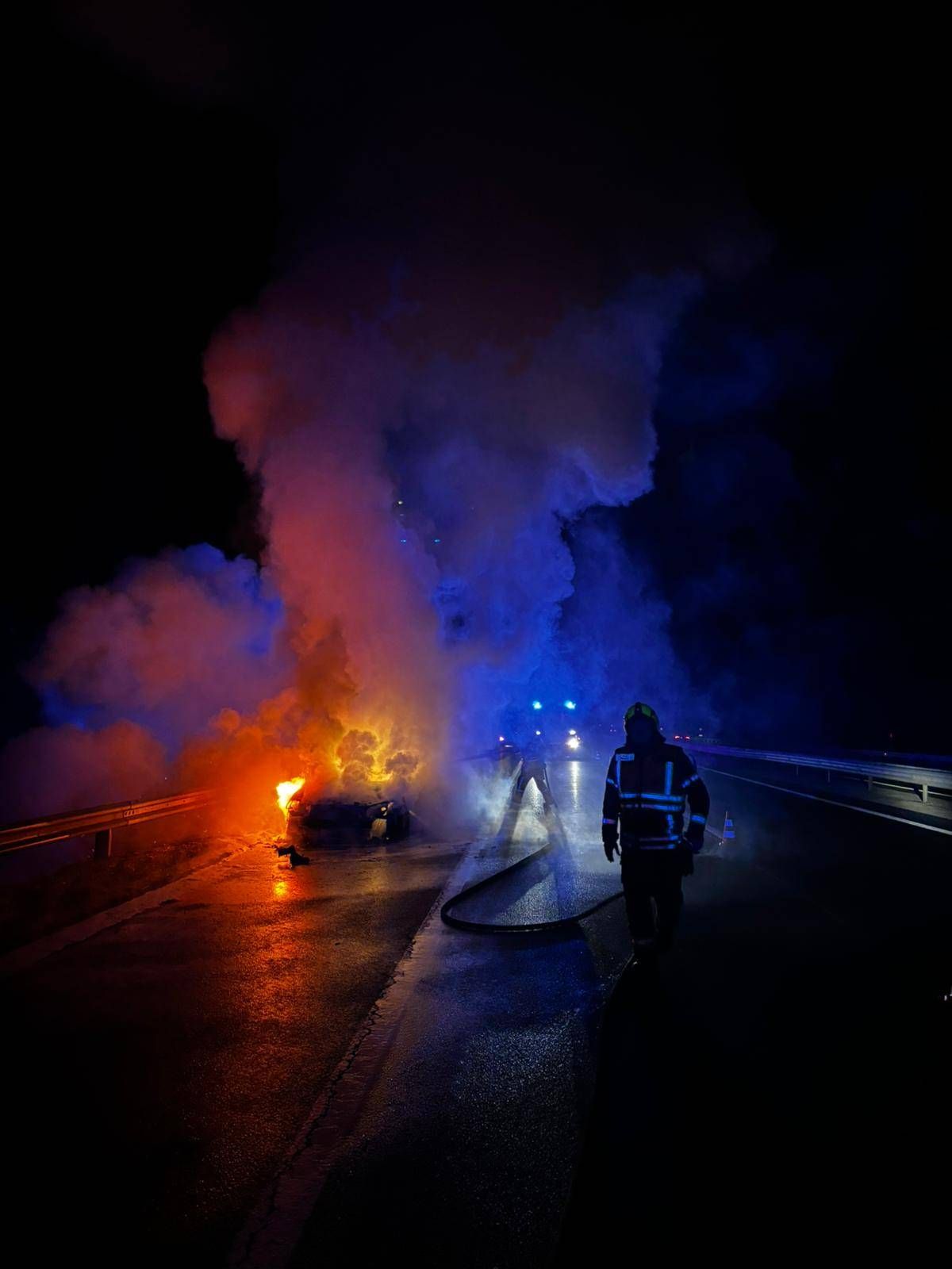 Ein Feuerwehrmann geht auf eine brennende Karre auf einer nassen Straße bei Nacht zu, mit Rauch und blinkenden Lichtern im Hintergrund.