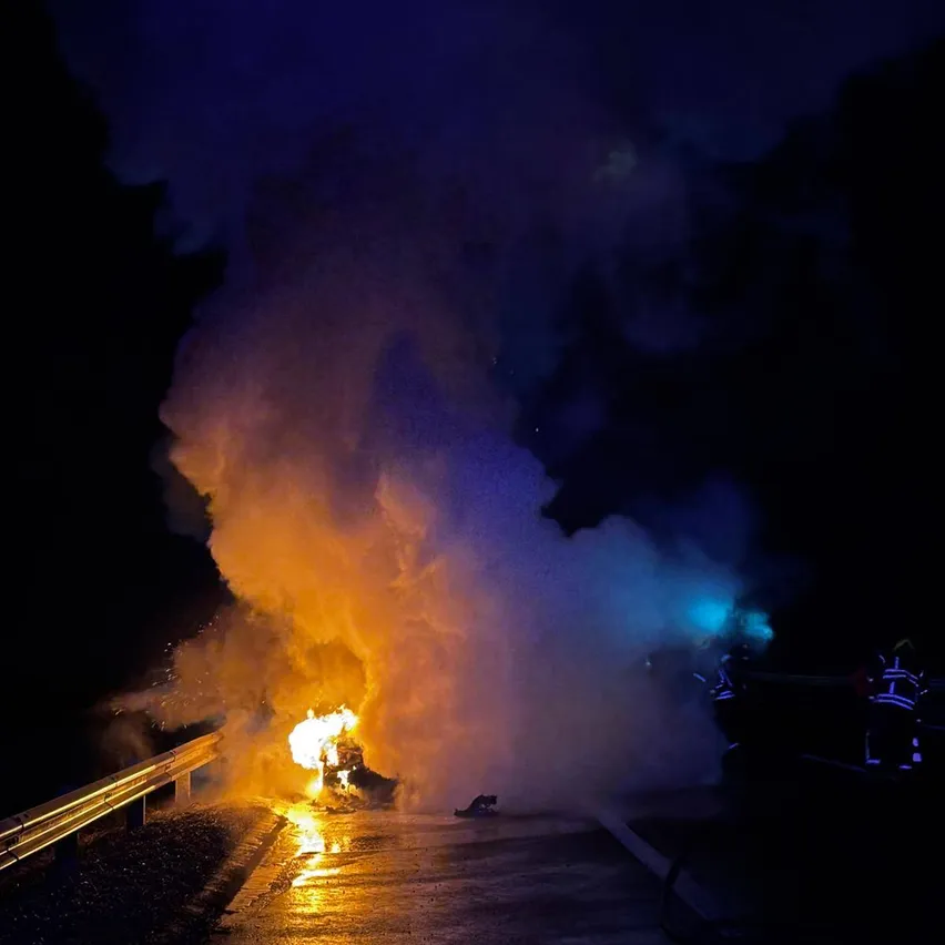 In der Nacht brennt ein Feuer auf einer nassen Straße, Feuerwehrleute in gelben Uniformen stehen in der Nähe, beleuchtet von blauen Lichtern.