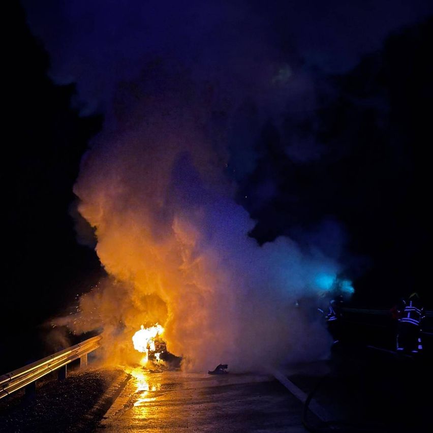 In der Nacht brennt ein Feuer auf einer nassen Straße, Feuerwehrleute in gelben Uniformen stehen in der Nähe, beleuchtet von blauen Lichtern.