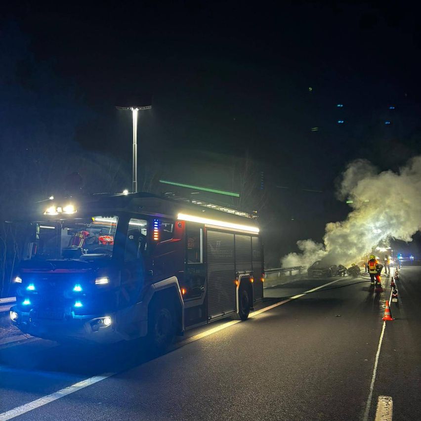Ein Feuerwehrwagen steht nachts mit eingeschalteten Lichtern auf der Straße. Rauch kommt von einem zerstörten Auto am Straßenrand. Eine Person in einem gelben Weste steht in der Nähe.