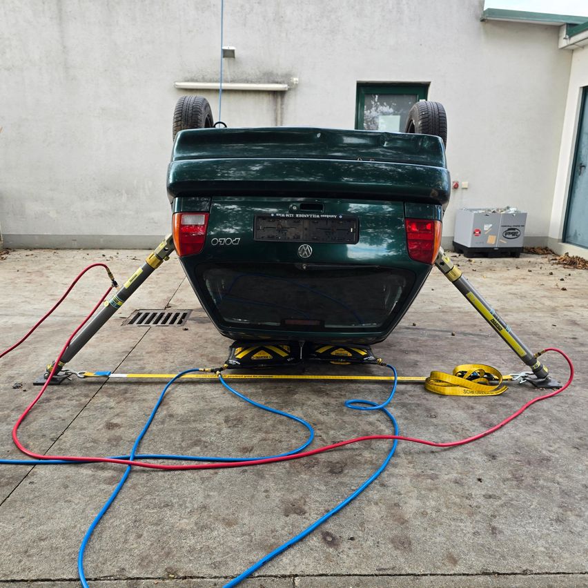Ein grünes Auto mit nach oben gerichteter Vorderseite wird von zwei Hebebügeln angehoben, die mit Kabeln verbunden sind, auf einem Betonboden.
