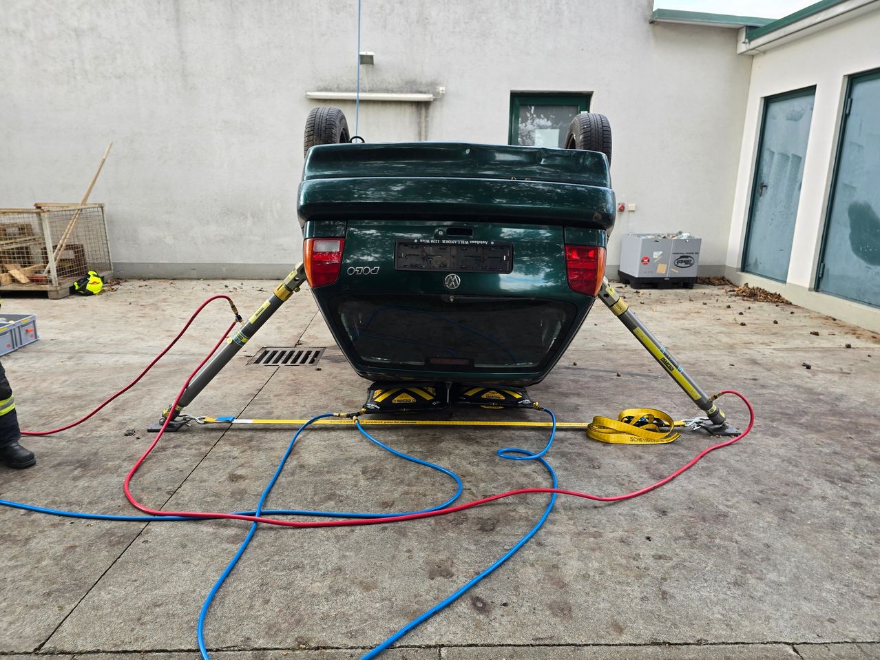 Ein grünes Auto mit nach oben gerichteter Vorderseite wird von zwei Hebebügeln angehoben, die mit Kabeln verbunden sind, auf einem Betonboden.