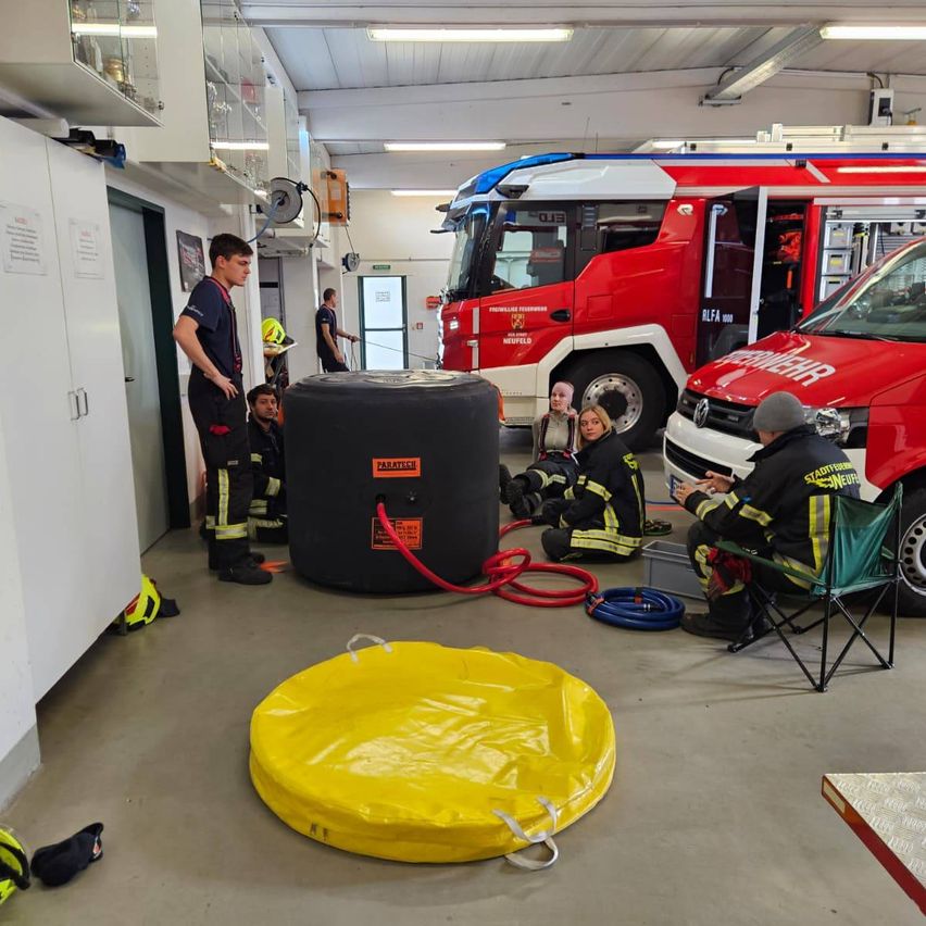 Feuerwehrleute versammeln sich um einen großen schwarzen Tank in einer Garage, wobei ein roter Feuerwehrwagen in der Nähe geparkt ist. Einige Leute sitzen auf Stühlen, während andere stehen. Auf dem Boden liegt eine gelbe aufblasbare Matte.