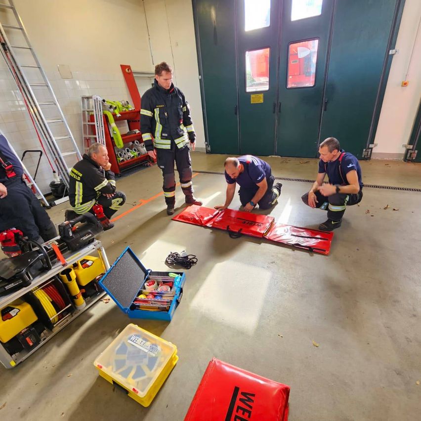 Feuerwehrleute üben in einem Raum mit roter und gelber Ausrüstung. Einer kniet, andere stehen. Werkzeuge und Ausrüstung liegen auf dem Boden und an der Wand.
