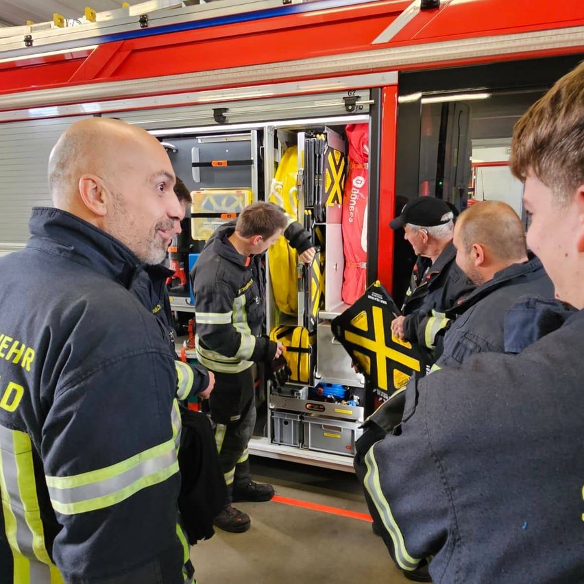 Mehrere Feuerwehrleute stehen vor einem Feuerwehrauto, einer hält eine offene gelbe Werkzeugkiste. Ein anderer Mann mit schwarzem Hut inspiziert die Ausrüstung.
