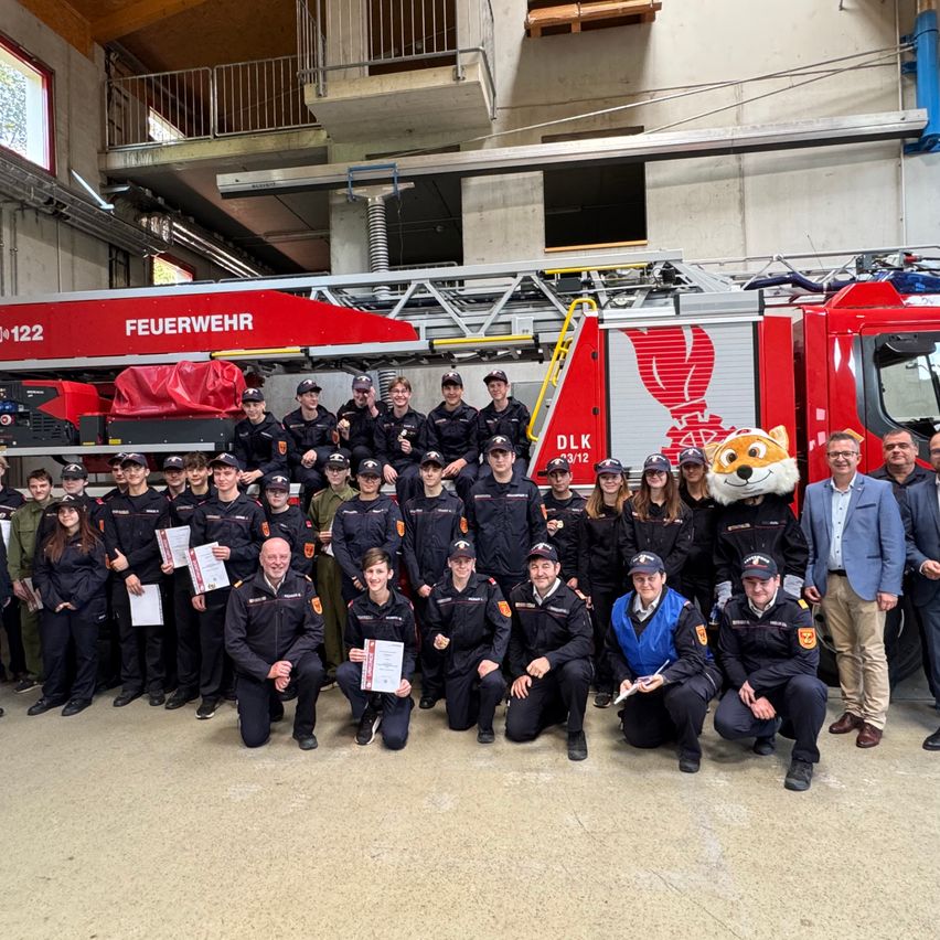 Eine Gruppe von Feuerwehrleuten und Beamten posiert für ein Foto vor einem roten Feuerwehrauto, mit einem Maskottchen und einigen, die Zertifikate in der Hand halten.