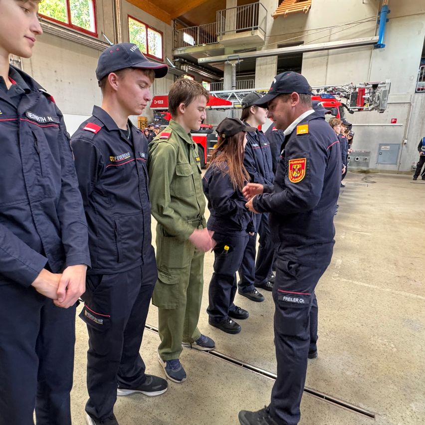 Eine Gruppe von Menschen in Uniformen steht in einer Garage. Sie unterhalten sich mit einem Mann in einer dunkelblauen Uniform. Der Mann trägt einen Hut und Turnschuhe.