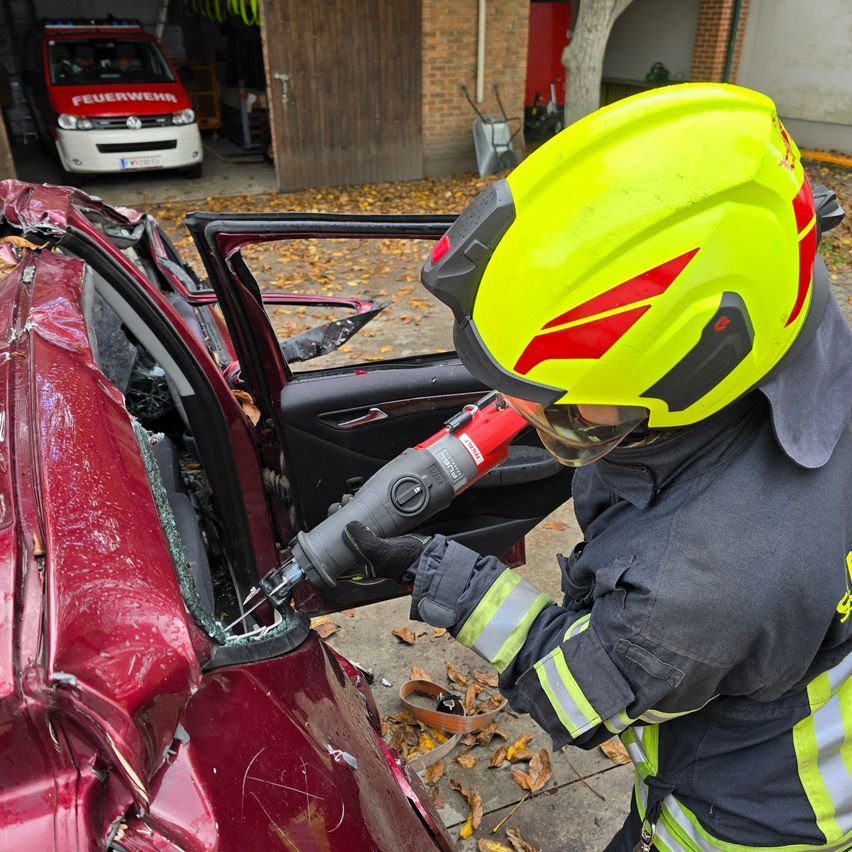 Ein Feuerwehrmann bricht mit einem Werkzeug ein Autofenster ein, während er einen gelben Helm trägt. Das Auto ist rot und beschädigt. Ein Feuerwehrwagen steht hinter dem Auto.