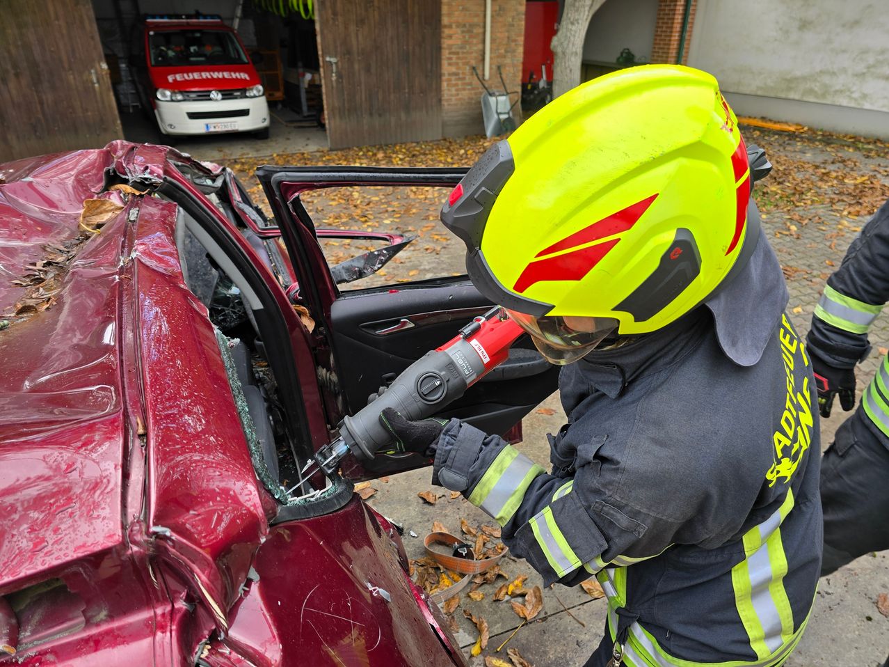 Ein Feuerwehrmann bricht mit einem Werkzeug ein Autofenster ein, während er einen gelben Helm trägt. Das Auto ist rot und beschädigt. Ein Feuerwehrwagen steht hinter dem Auto.