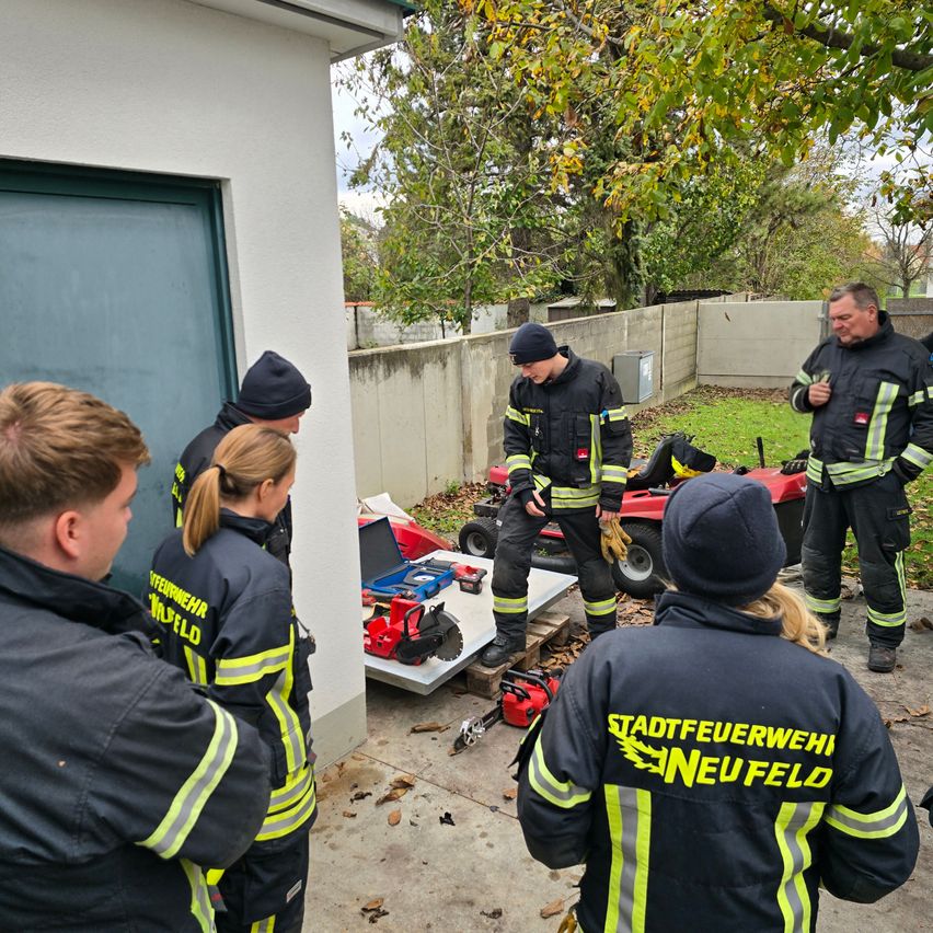 Feuerwehrleute in Uniform stehen auf einem Hof. Sie stehen um eine geschlossene Garagentür herum. Dahinter ist ein kleines rotes Fahrzeug geparkt.