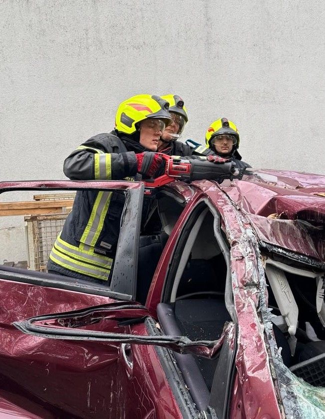 Drei Feuerwehrleute in gelben und schwarzen Uniformen und Helmen arbeiten an einem beschädigten roten Auto. Sie verwenden Werkzeuge, um Teile des Autos zu entfernen.