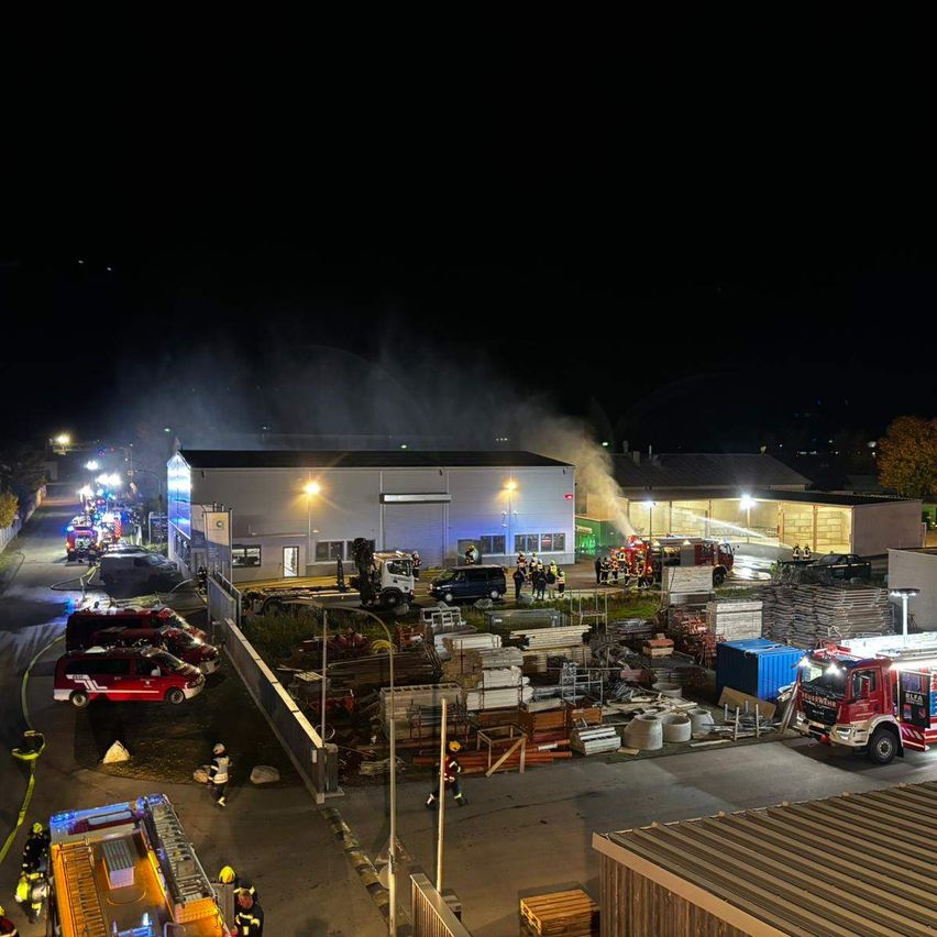 Eine Nachtszene mit mehreren Feuerwehrfahrzeugen und Personal um ein brennendes Gebäude. Das Gebiet ist gut beleuchtet.