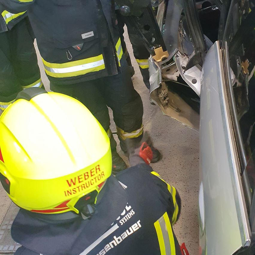 Ein Feuerwehr-Instruktor gibt in einem Auto Unterricht, ein helmtragender Instruktor kniet neben einer beschädigten Fahrzeugtür.