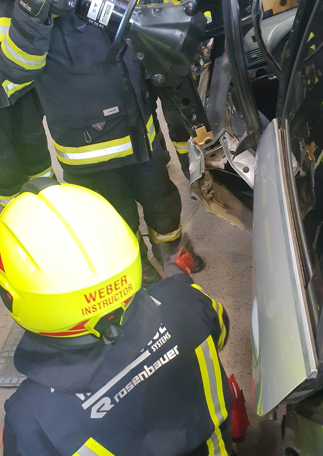 Ein Feuerwehr-Instruktor gibt in einem Auto Unterricht, ein helmtragender Instruktor kniet neben einer beschädigten Fahrzeugtür.