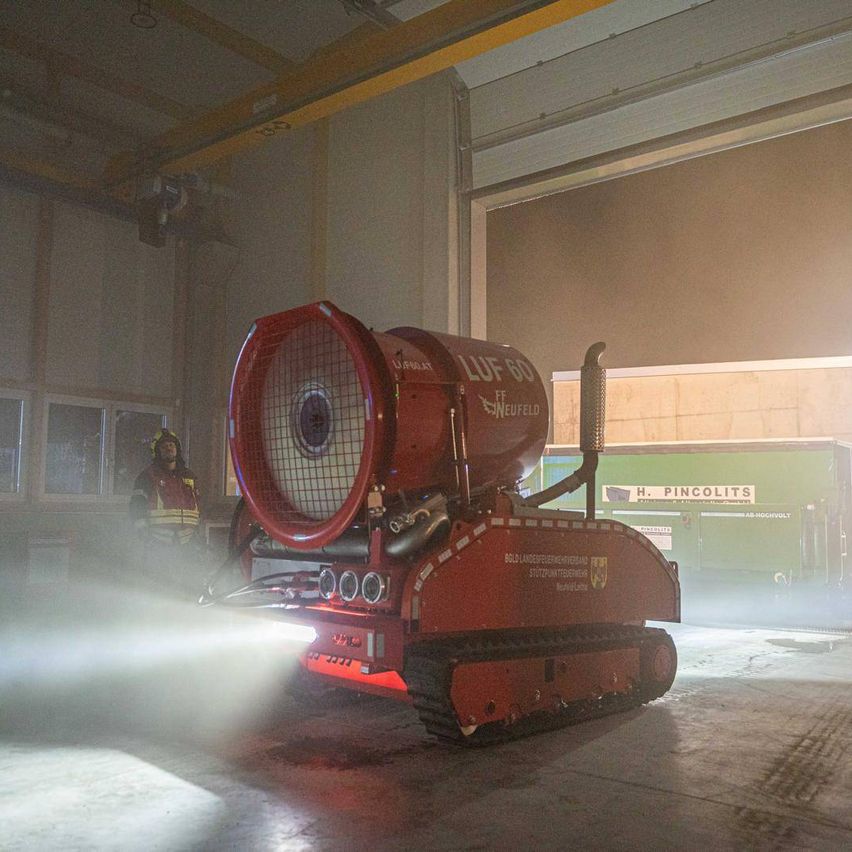 Ein Feuerwehrmann steht neben einer großen roten Maschine mit 'LUF 60 FF' darauf, die Wasser in einem Lager sprüht. Die Szene ist schwach beleuchtet.