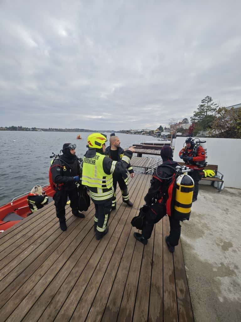 Eine Gruppe von Tauchern mit Sauerstoffflaschen bereitet sich darauf vor, in ein Gewässer zu tauchen, wobei ein Boot und ein Steg im Hintergrund zu sehen sind.