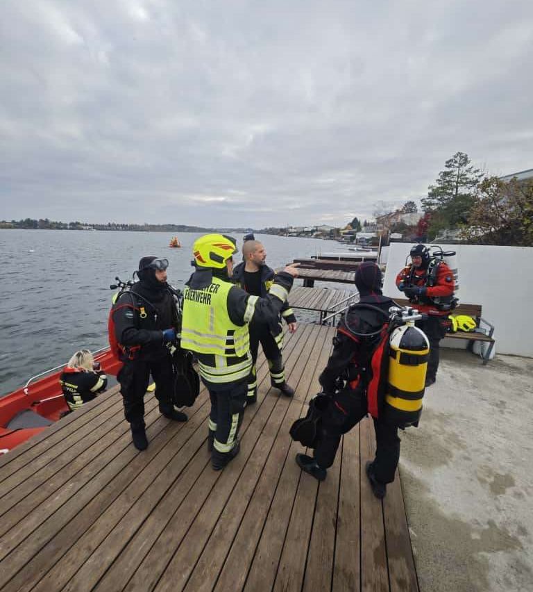Eine Gruppe von Tauchern mit Sauerstoffflaschen bereitet sich darauf vor, in ein Gewässer zu tauchen, wobei ein Boot und ein Steg im Hintergrund zu sehen sind.