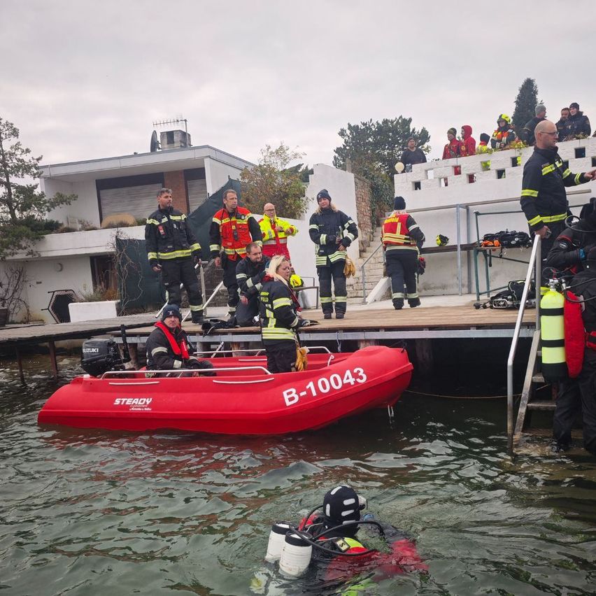 Ein Rettungsteam bereitet sich darauf vor, einem Taucher in Not bei einem roten Boot mit der Bezeichnung B-10043 zu helfen. Feuerwehrleute in gelben und schwarzen Uniformen stehen auf einem Steg.