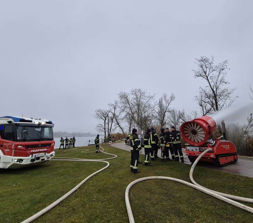 Ein Feuerwehrfahrzeug mit angeschlossenem Schlauch steht auf einer Rasenfläche in der Nähe eines Sees. Feuerwehrleute stehen um ein rotes Gerät herum, das Wasser sprüht.