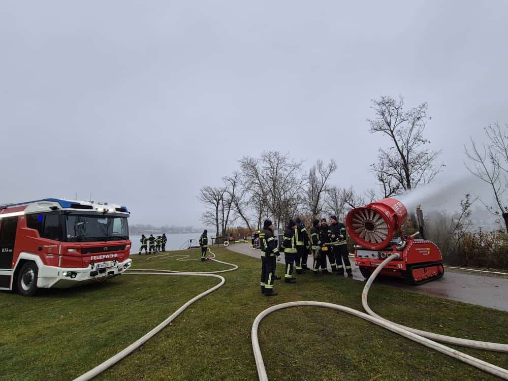 Ein Feuerwehrfahrzeug mit angeschlossenem Schlauch steht auf einer Rasenfläche in der Nähe eines Sees. Feuerwehrleute stehen um ein rotes Gerät herum, das Wasser sprüht.