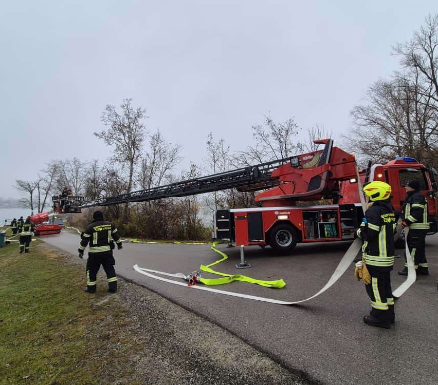 Feuerwehrleute bereiten einen Feuerwehrwagen auf einer Straße mit Bäumen und einem See im Hintergrund vor.